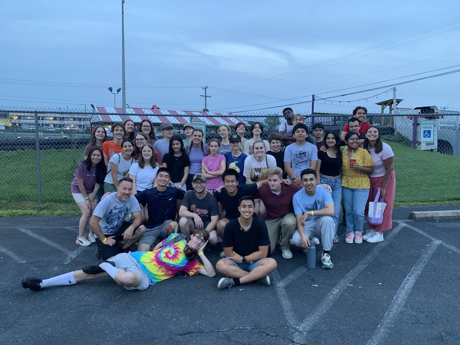 A large group of youth and leaders gathered for a photo in a parking lot at dusk